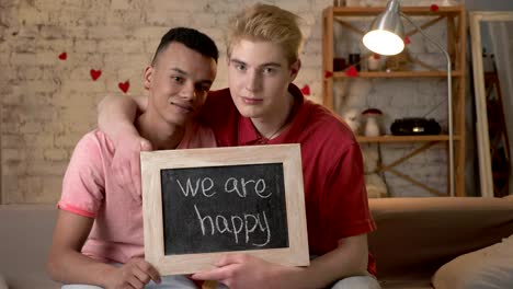 A-sad-international-gay-couple-is-sitting-on-the-couch-and-holding-a-sign.-We-are-not-happy.-Look-at-the-camera.-Home-comfort-on-the-background.-60-fps