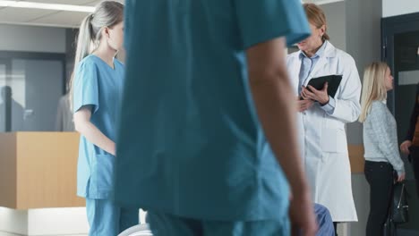 In-the-Hospital-Lobby,-Nurse-Pushes-Elderly-Patient-in-the-Wheelchair,-Doctor-Talks-to-Them-while-Using-Tablet-Computer.-Clean,-New-Hospital-with-Professional-Medical-Personnel.-Slow-Motion.