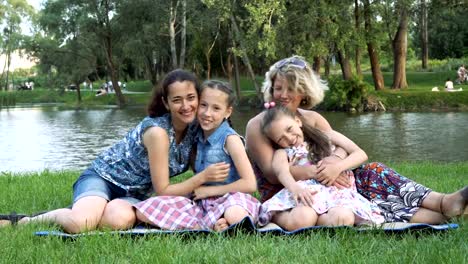 Familia-mujer-feliz-jugando-en-el-parque.-Dos-madres-y-dos-hijas-se-sientan-en-el-césped-junto-al-río-en-el-atardecer-riendo,-abrazando-y-besando.