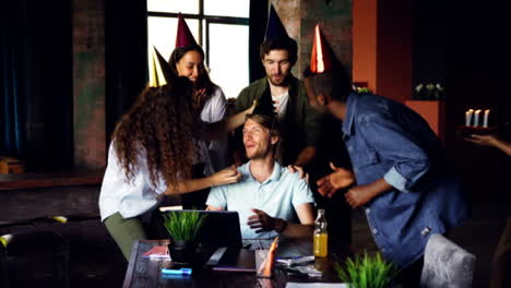 Company-employees-are-congratulating-their-boss-on-birthday-bringing-cake-and-party-hats,-young-man-is-blowing-candles-and-doing-high-five-while-workers-are-clapping-hands.