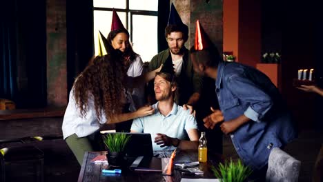 Company-employees-are-congratulating-their-boss-on-birthday-bringing-cake-and-party-hats,-young-man-is-blowing-candles-and-doing-high-five-while-workers-are-clapping-hands.