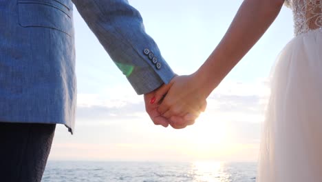 Unrecognizable-wedding-couple-holding-hands-on-sunset-or-sunrise-background.-Bride-and-groom-standing-outdoor-near-sea.-Sun-flares.-Slow-motion