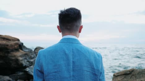 Portrait-of-young-attractive-business-man-or-groom-stands-on-sea-shore-with-rocks-and-watching-beautiful-sunset-or-sunrise-alone.-4k.-Slow-motion