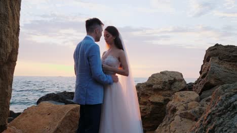 Beautiful-young-wedding-couple-standing-on-sea-shore-with-rocks.-Newlyweds-spend-time-together:-embrace,-kiss-and-care-for-each-other.-Love-concept.-Bride-smiling-to-groom