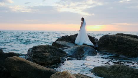 Girl-in-wedding-luxury-dress-posing-on-sea-shore.-Bride-on-a-rocks.-Beautiful-waves-near-to-her.-Woman-enjoying-happy-moments-with-nature.-Sunrise-or-sunset-time.-Slow-motion