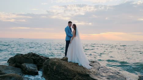 Beautiful-young-wedding-couple-standing-on-sea-shore-with-rocks.-Newlyweds-spend-time-together:-embrace,-kiss-and-care-for-each-other.-Love-concept