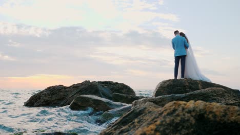 Hermosa-joven-pareja-de-pie-en-la-orilla-del-mar-con-las-rocas.-Recién-casados-pasan-tiempo-juntos:-abrazar,-besar-y-cuidar-de-uno-al-otro.-Concepto-de-amor