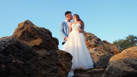 Beautiful-young-wedding-couple-standing-on-sea-shore-with-rocks.-Newlyweds-spend-time-together:-embrace,-kiss-and-care-for-each-other.-Camera-moves-from-behind-rock