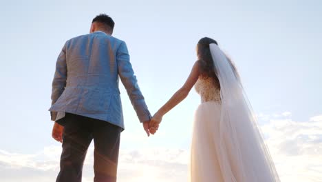 Unrecognizable-wedding-couple-holding-hands-on-sunset-or-sunrise-background.-Bride-and-groom-standing-outdoor-near-sea.-Sun-flares.-Slow-motion