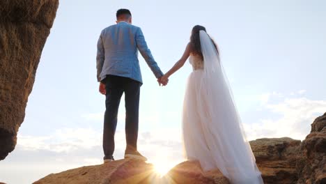 Novios-irreconocible-manteniendo-las-manos-sobre-fondo-de-atardecer-o-amanecer.-Novia-y-el-novio-de-pie-al-aire-libre-junto-a-la-mar.-Las-llamaradas-del-sol.-Cámara-lenta