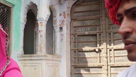 Pan-close-up-from-a-handsome-man-with-red-turban-to-a-pretty-lady-in-pink-sari-talking-in-front-of-a-decorative-entry-old-haveli-building