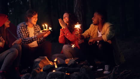 Young-woman-is-celebrating-birthday-in-forest-with-friends-blowing-candles-on-cake-and-clapping-hands-while-people-around-are-holding-bengal-lights-and-congratulating-her.