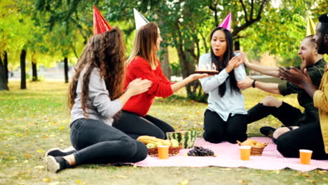 Friends-are-congratulating-Asian-woman-on-birthday-giving-cake-making-surprise,-girl-is-blowing-candles,-smiling-and-clapping-hands-during-outdoor-party-in-park.