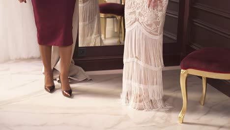 A-charming-young-blonde-in-an-embroidered-fancy-dress-with-fringe-at-the-bottom-is-turning-around-in-front-of-the-mirror-in-an-atelier-and-the-designer-is-looking-at-it.