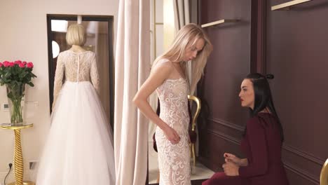 A-designer-in-a-dark-red-dress-is-sitting-on-the-chair-and-checking-how-the-embroidered-fancy-dress-fits-her-blonde-client,-who-stands-in-front-of-her.-She-is-looking-at-the-cloth-examining-it.