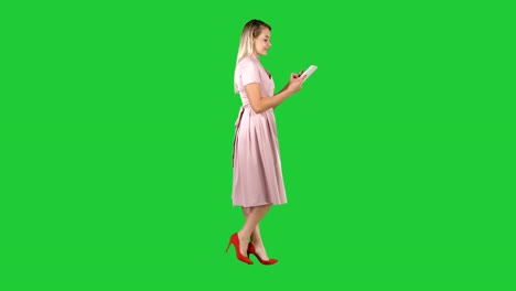 Blonde-in-pink-dress-walking-and-using-tablet-on-a-Green-Screen,-Chroma-Key