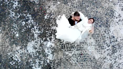 invierno-de-la-boda.-Vista-aérea-de-la-pareja-de-recién-casados-en-vestidos-de-novia-están-bailando-la-danza-de-la-boda-en-un-parque-cubierto-de-nieve,-sobre-el-trasfondo-de-adoquines