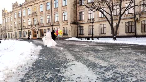 invierno-de-la-boda.-Vista-aérea,-pareja-de-recién-casados-en-vestidos-de-novia-es-runing-con-globos-de-colores-por-el-Parque-cubierto-de-nieve,-en-el-contexto-de-la-arquitectura-antigua-y-piedras