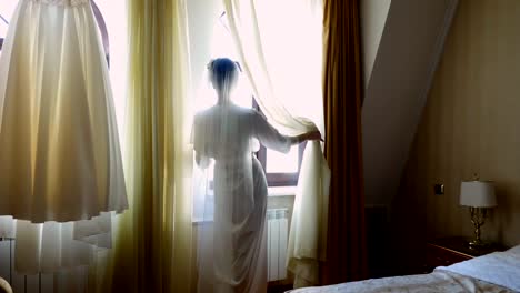 a-bride,-beautiful-girl,-in-veil-and-white-peignoir,-robe,-looks-out-the-window.-Morning-of-the-bride