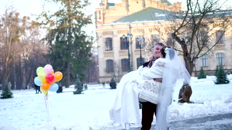 winter-wedding.-newlywed-couple-in-wedding-dresses.-groom-holds-bride-in-his-arms,-spinning.-they-are-happy,-smiling-to-each-other.-background-of-ancient-architecture,-snow-covered-park