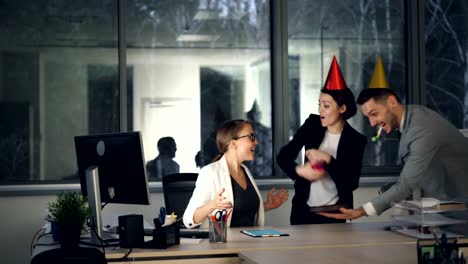 Bonita-rubia-está-trabajando-en-la-oficina-moderna-cuando-sus-colegas-están-trayendo-la-torta-de-cumpleaños,-gorros-de-fiesta-y-presente,-chica-es-abrir-la-caja-de-regalo-y-disfrutar-Felicidades