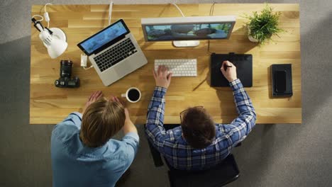 Top-View-of-Two-Young-Talented-Photographers-Discussing-Footage.-One-of-Them-Works-on-a-Digital-Drawing-Tablet-at-His-Desktop-Computer.-Processing-Wedding-Photography.-His-Worplace-is-Covered-with-Sunlight.