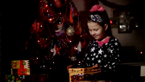 In-der-Dämmerung-der-Nacht,-eine-hübsche-blonde-Mädchen-mit-rosa-Schleife-im-Haar,-in-einem-ein-schönes-Kleid-bewundert-Weihnachtsgeschenke,-öffnet-sich-eine-große-Kiste,-erfüllt-den-Raum-mit-einem-magischen-Glanz,-sie-ist-glücklich,-froh.-Im-Hintergrund-ist-ein-Weihnachtsbaum-in-Farbe
