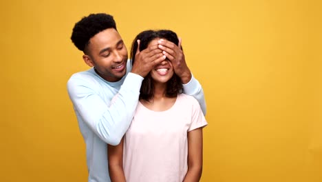 Young-smiling-african-man-closing-his-girlfriends-eyes-and-hugging-her-isolated-over-yellow-background