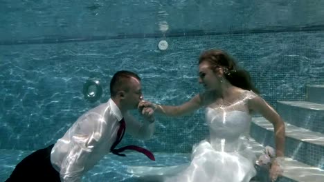 Happy-groom-and-bride-in-white-wedding-dress-sitting-underwater-on-the-bottom-of-the-pool.-The-bridegroom-floats-and-kisses-the-bride's-hand