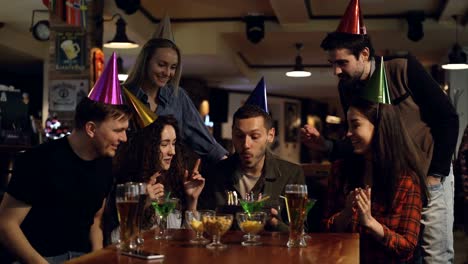 Handsome-young-man-is-making-wish-and-blowing-out-candles-on-cake-while-celebrating-birthday-in-bar-with-friends.-Happy-people-are-congratulating-him-and-clapping-hands.