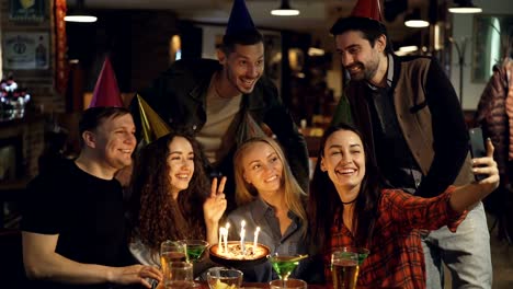 Happy-attractive-students-in-birthday-party-hats-are-recording-video-with-smartphone-sitting-at-table-in-bar.-They-are-posing-with-cake,-talking-and-laughing.