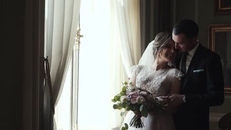 Marrying-couple-embrace-and-kiss-standing-near-huge-window-in-room