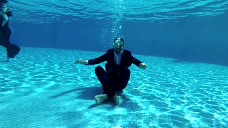 The-happy-groom-in-a-suit-and-a-white-shirt-goes-down-underwater-to-the-bottom-of-the-pool.-He-sits-at-the-bottom-in-a-Lotus-position,-looks-at-the-camera-and-smiles.-Slow-motion.-Action-camera-underwater.