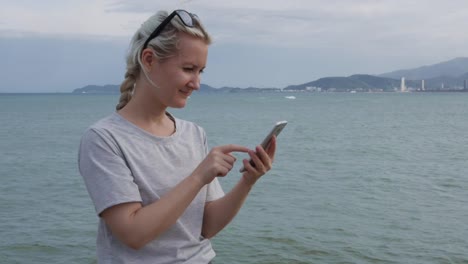 Schöne-schlanke-Frau-mit-einem-Zopf-Blondine-in-einem-grauen-T-shirt-stehend-und-mit-Smartphone-über-Hintergrund-Meer-und-Inseln.-Bildschirm-berühren-Mädchen