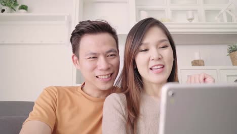 Asian-paar-mit-Tablet-Videoanruf-mit-Freund-im-Wohnzimmer-zu-Hause,-süßes-Paar-Liebe-Moment-beim-liegen-auf-dem-Sofa,-wenn-zu-Hause-entspannt-genießen.-Lifestyle-paar-entspannen-zu-Hause-Konzept.