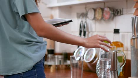 Glückliche-junge-asiatische-Frau,-die-eine-Flasche-Wasser-in-ein-Glas-und-mit-Tablette.-Technik,-soziales-Netzwerk,-Kommunikationskonzept-in-der-Küche-zu-Hause.