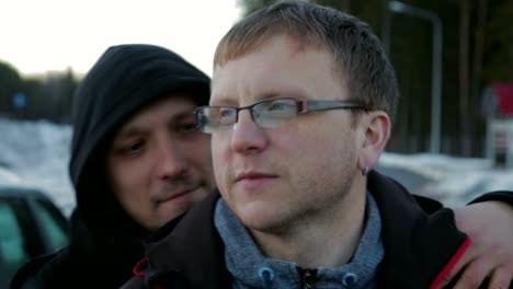 Two-LGBT-guys,-bald-with-a-beard-and-wearing-glasses-and-a-hat-hugging-and-chatting-on-the-side-of-the-road-near-the-car