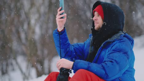 Männliche-Snowboarder-sitzen-auf-dem-Schnee-und-machen-Fotos-am-Telefon-schöne-Landschaft-des-Resorts-für-soziale-Netzwerke