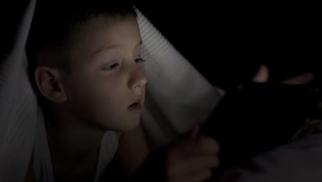 A-happy-boy-lies-in-bed-under-a-blanket-and-plays-on-a-tablet-in-a-game-in-the-dark.-The-face-of-the-child-is-lit-by-a-bright-monitor