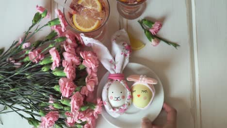 Die-weibliche-Hand-legt-Teller-mit-Osterei,-das-für-Osterhasen-und-Osterschick-geschmückt-ist,-vor-dem-Hintergrund-rosa-Nelken-und-Limonade