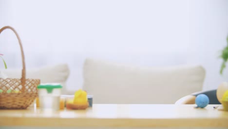 Young-adorable-boy-is-hiding-under-the-table-full-of-Easter-decorations-and-is-playing-with-Easter-bunny-in-his-hand.-Bunny-theatre.-Discussion.-Conversation:-cheerful-boy-and-a-soft-bunny.