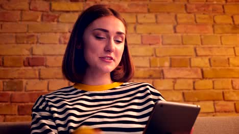 Closeup-portrait-of-young-pretty-girl-having-a-video-call-on-the-tablet-and-talking-cheerfully-sitting-on-the-sofa-in-a-cozy-apartment-indoors