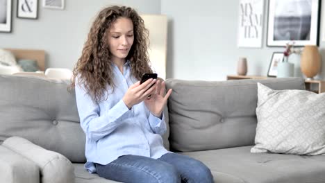 Junge-lockige-Haar-Frau-mit-Smartphone-beim-Entspannen-auf-dem-Sofa