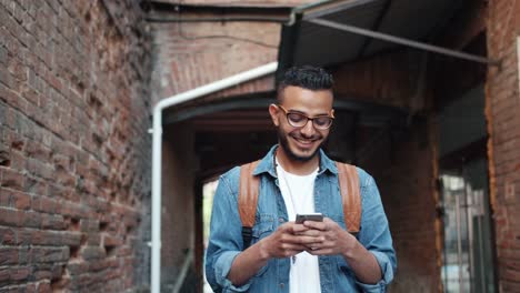 Guapo-árabe-barbudo-disfrutando-de-la-aplicación-de-teléfono-inteligente-moderno-sonriendo-al-aire-libre