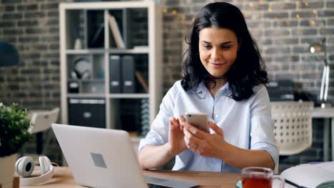 Hübsches-Mädchen-berührt-Smartphone-Bildschirm-und-lächelnd-sitzen-am-Schreibtisch-im-Büro