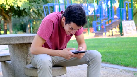 Joven-usando-su-teléfono-inteligente-sentado-en-un-banco-del-parque