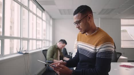 Geschickter-junger-afrikanischer-Geschäftsmann-mit-Blick-auf-Kamera-hält-digitales-Tablet-im-Büro-mit-Team-Kollegen-im-Hintergrund