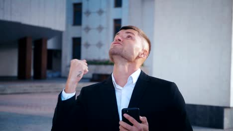 Businessman-celebrating-success.-Man-received-a-message-with-good-news.-Successful-Businessman-showing-Yes-gesture,-extremely-happy-about-good-news-from-smartphone.-Office-building-background.