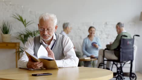 Rack-Fokus-der-Gruppe-von-drei-älteren-Menschen,-zwei-Seniorinnen-und-behinderten-alten-Mann-im-Rollstuhl,-Karten-im-Pflegeheim-spielen.-Intelligenter-alter-Mann-in-Brillen-lese-Buch-am-Tisch-im-Vordergrund