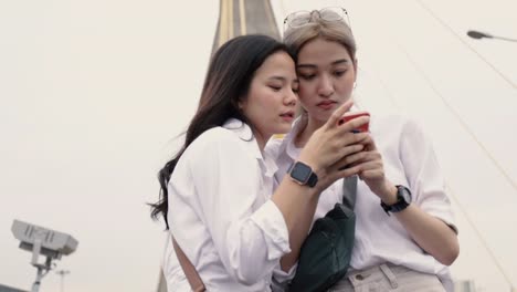 Asian-lesbian-couples-using-smartphones-browsing-the-website-while-standing-on-the-bridge.-Low-angle-shot.